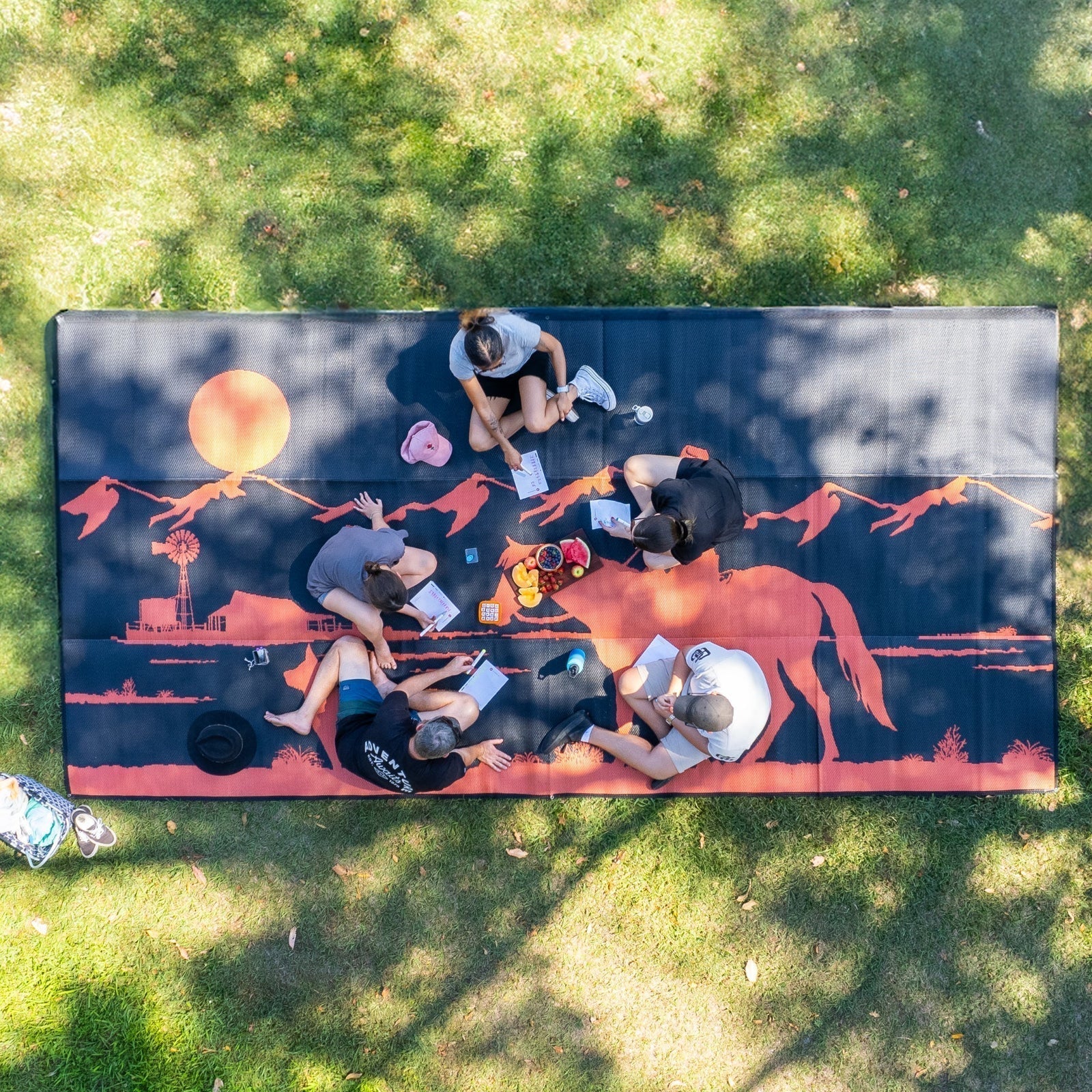 Five friends sitting on a picnic mat, enjoying food and drinks together in a cheerful outdoor setting.